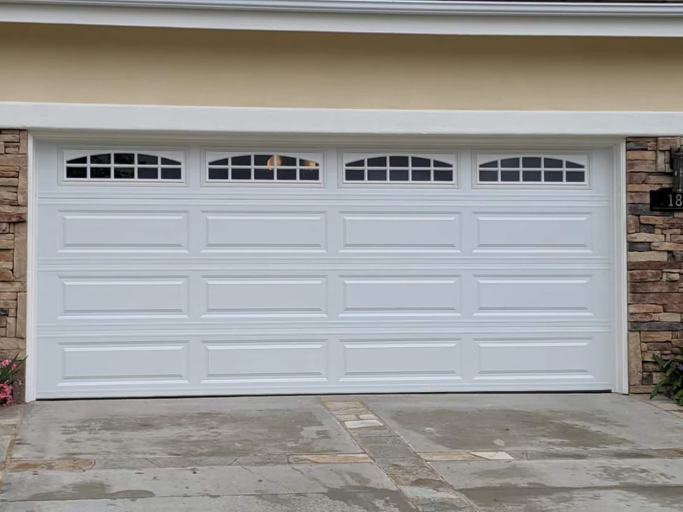 White two-car garage door with four arched window inserts featuring grilles, and raised rectangular panels, on a house with stone veneer siding.