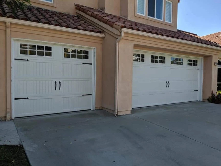 Two white carriage house style garage doors (one single, one double) with black decorative hardware and windows, on a stucco house with a tile roof.