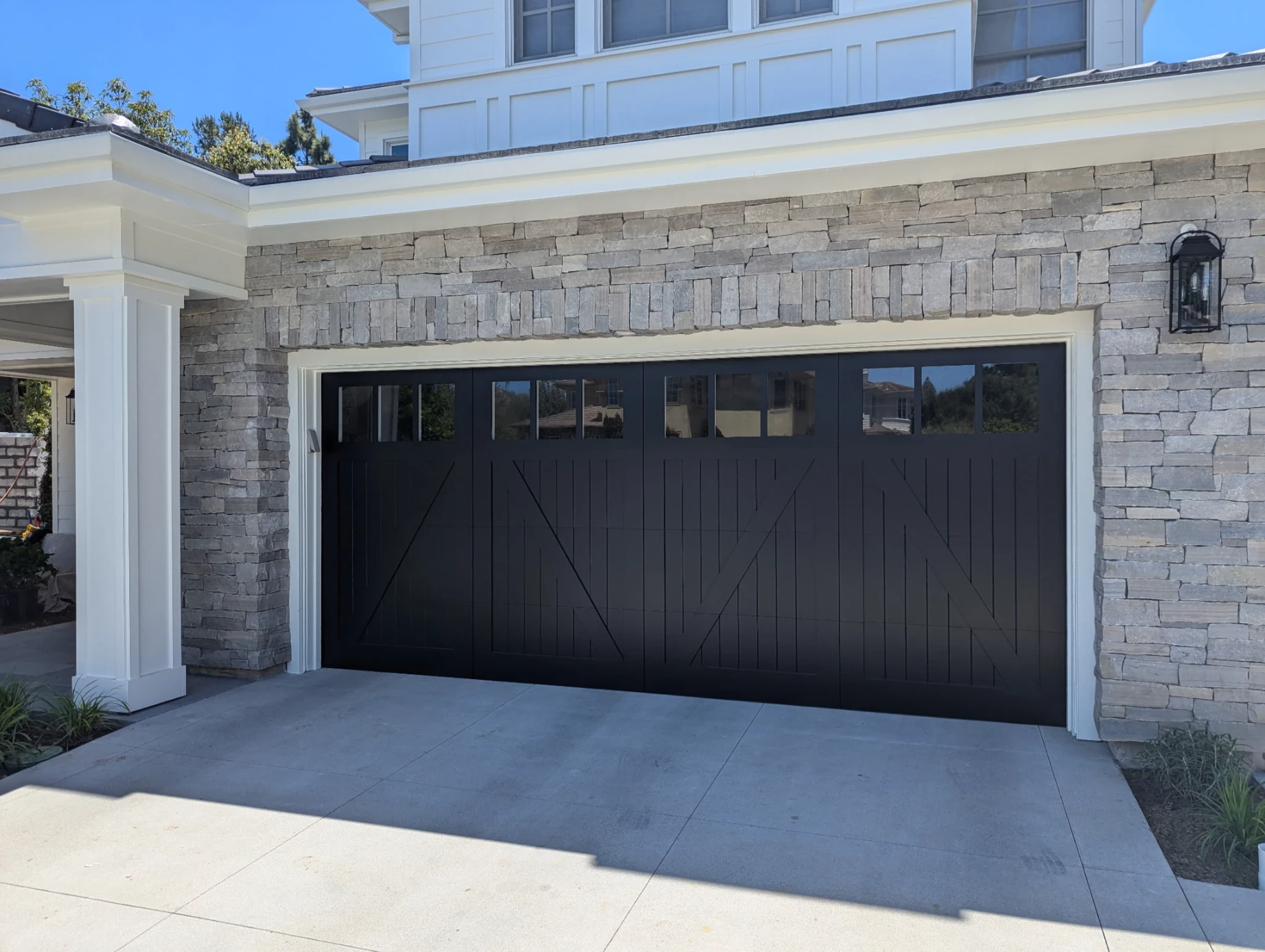 Black farmhouse-style two-car garage door with cross-buck "X" panel design and a row of rectangular windows, on a house with light grey stone veneer.