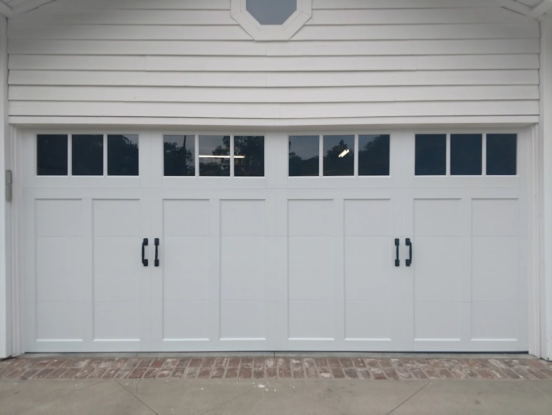 White two-car garage door with black decorative handles and a row of rectangular windows across the top, on a house with white lap siding.